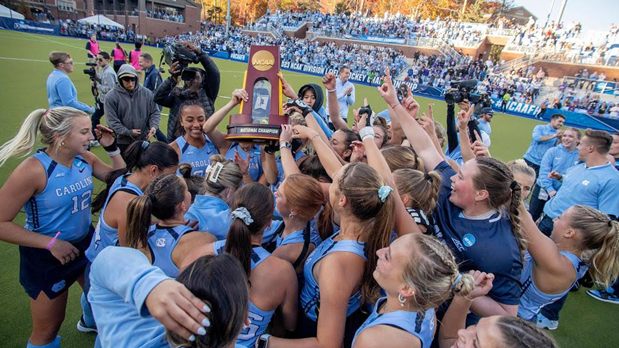 Coach Matson and UNC Field Hockey team in carolina blue uniforms celebrating with NCAA National Champion trophy on field.